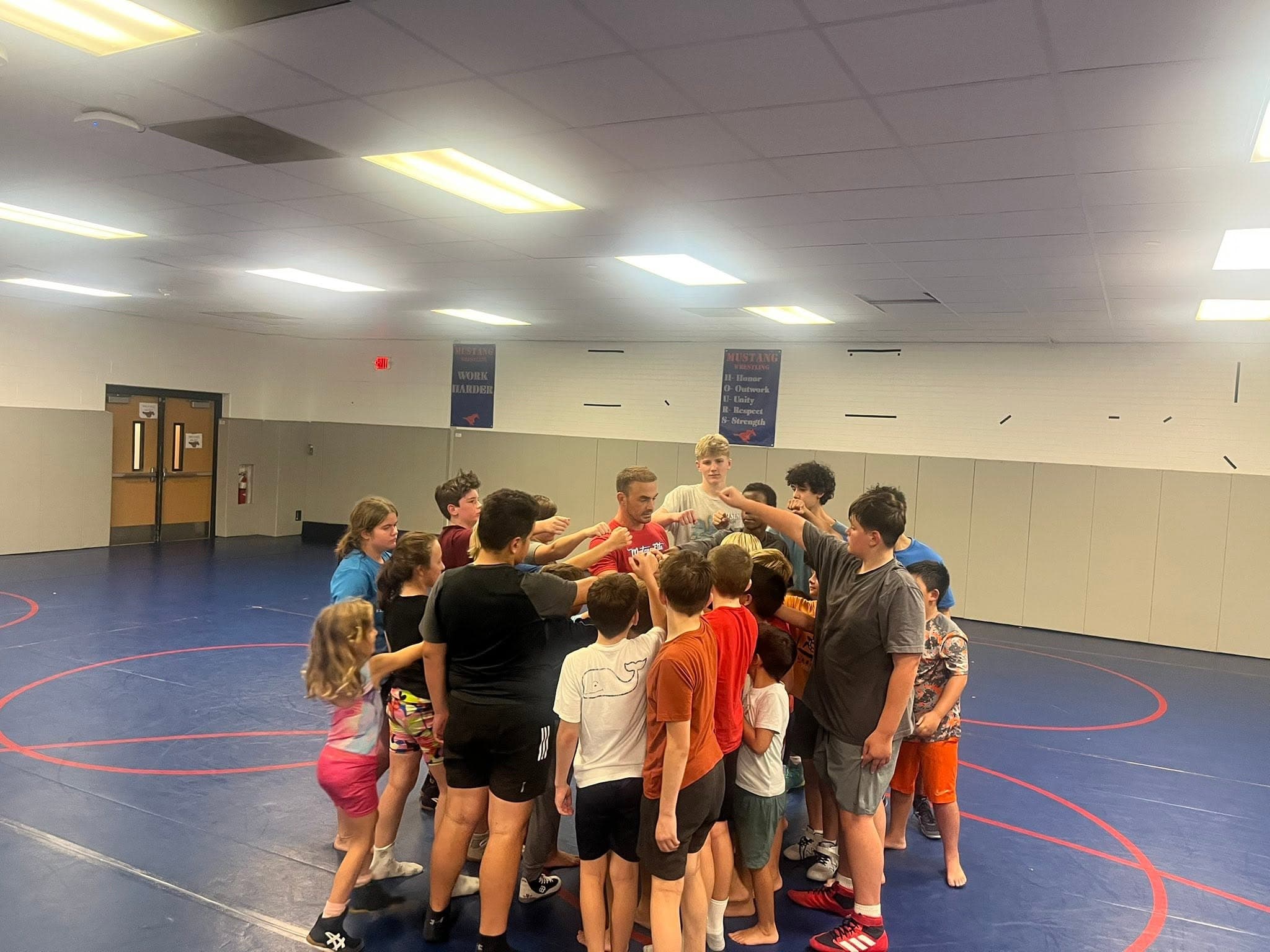 Coach leading a team huddle with wrestlers of all ages on the mat